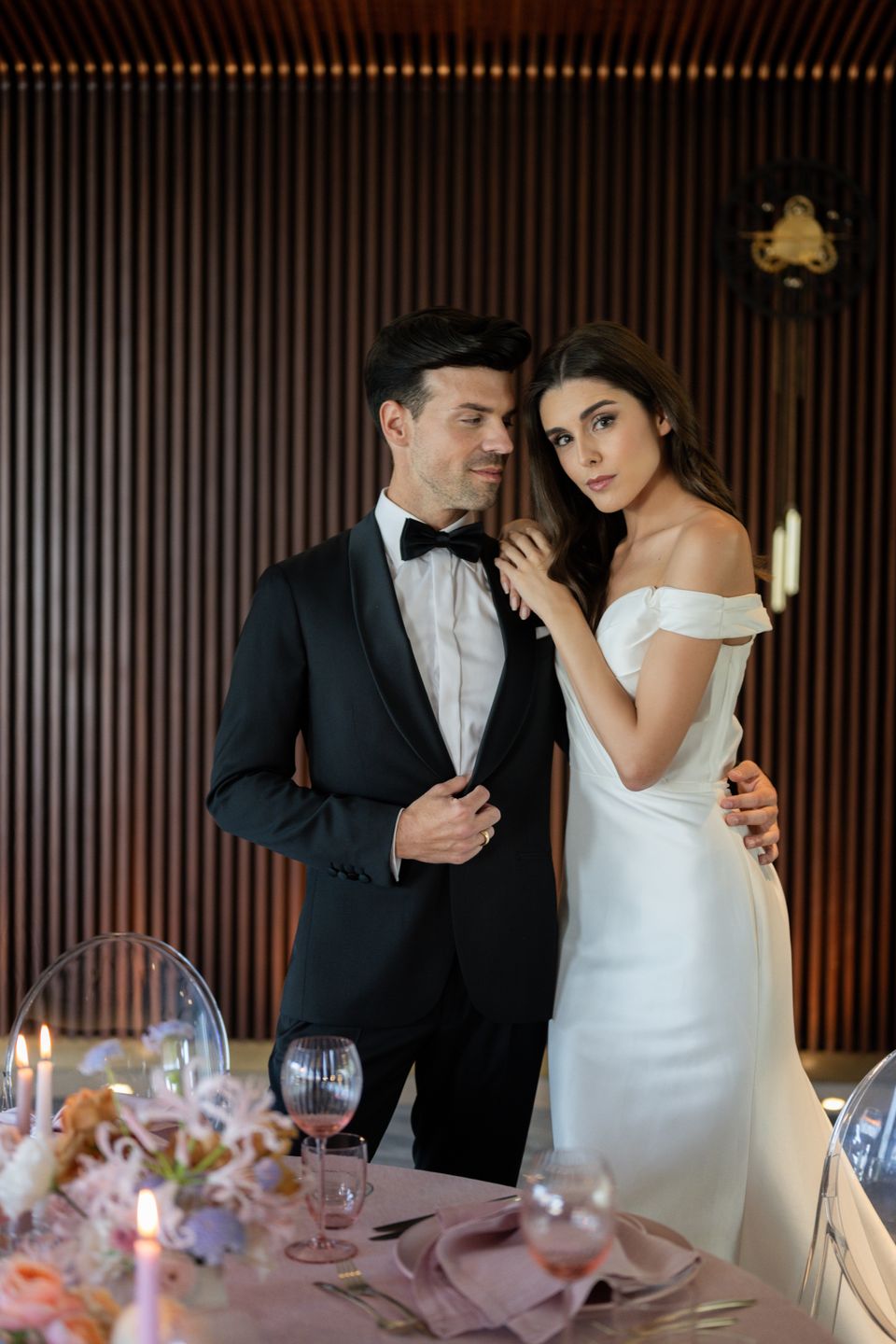 Vivienne Oesch and Vasilis Zorba posing as bride and groom at a wedding table. She wears a a white, off-the-shoulders dress, he wears a tuxedo. Bridal glam makeup. Hair and makeup by Denise Dedich.