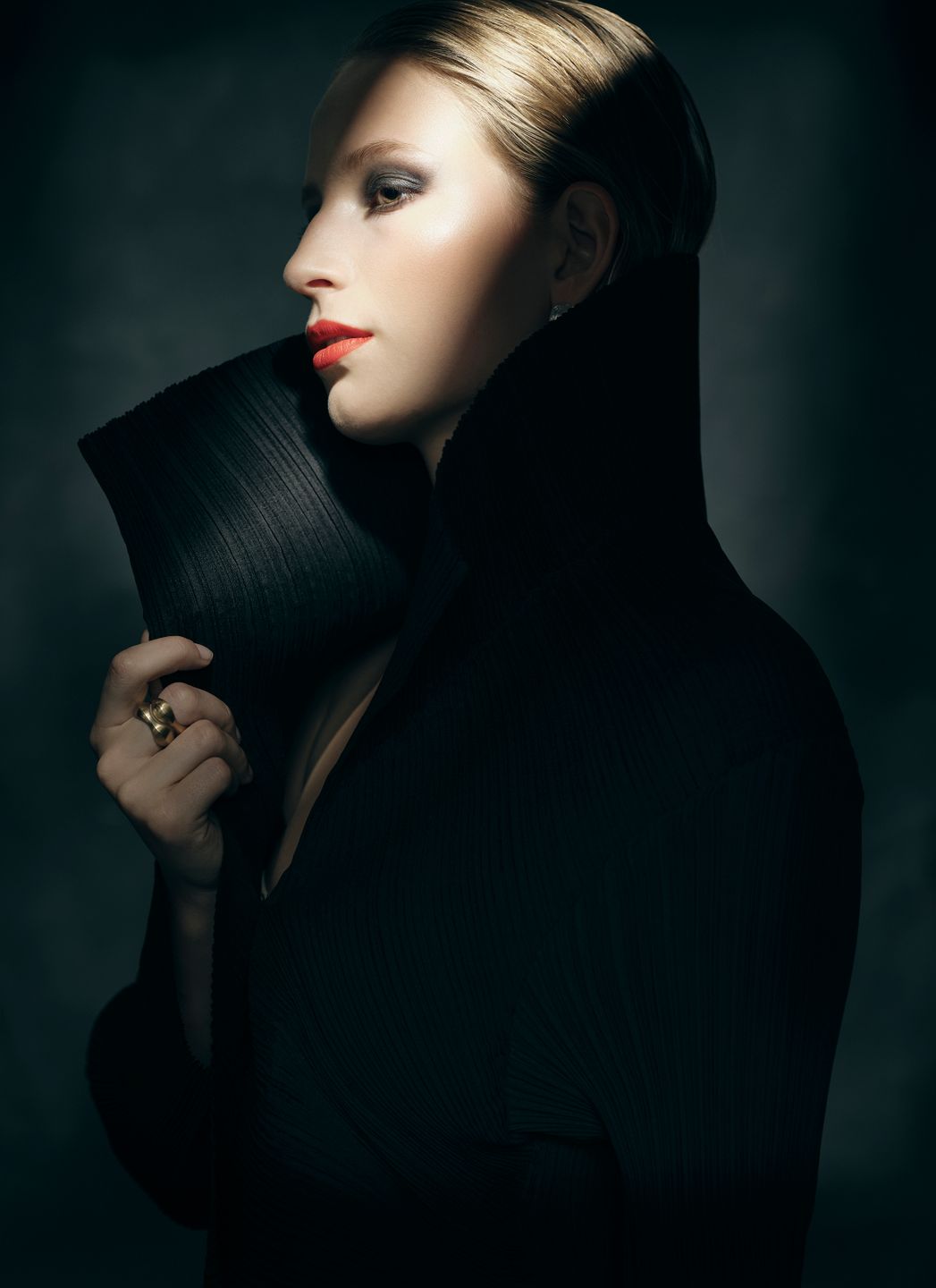 Model Joy Emmenegger in a black coat posing in front of a dark background. Red lipstick, glam makeup. Hair and makeup by Denise Dedich.