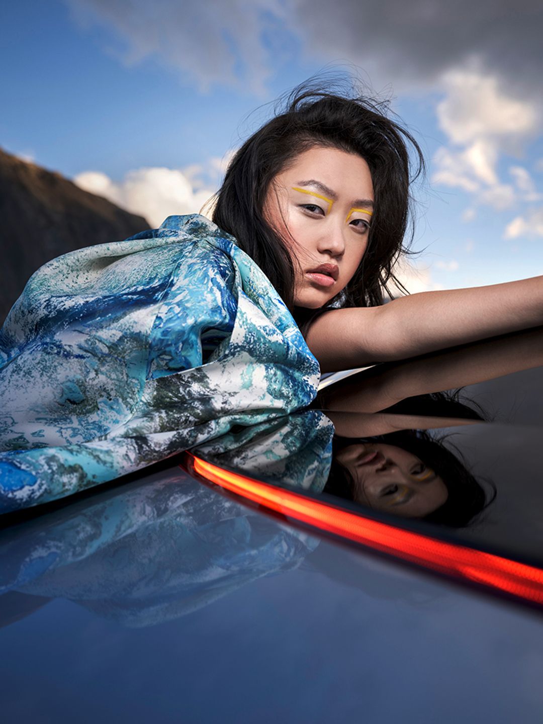 Model Dami J leans over a Mercedes EQE. She has a bold yellow graphic liner for makeup. Editorial makeup.  Hair and makeup by Denise Dedich.