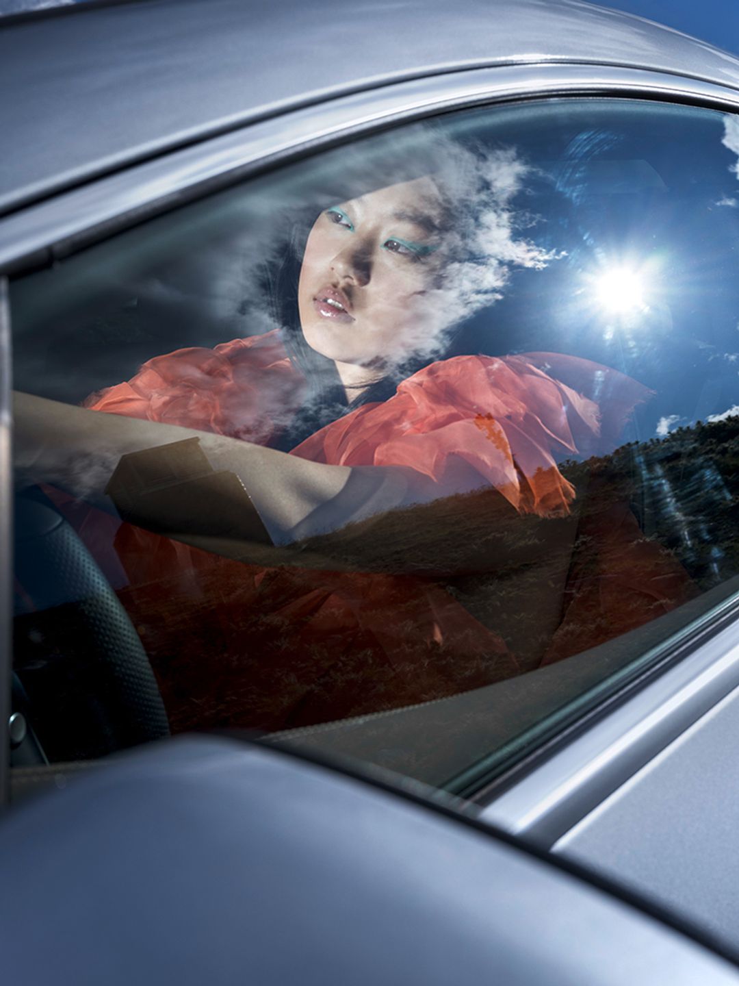 Model Dami J in a Mercedes EQE looking out the window. She wears an Amorphose orange dress. She has a turquoise graphic liner for makeup. Editorial makeup. Hair and makeup by Denise Dedich.