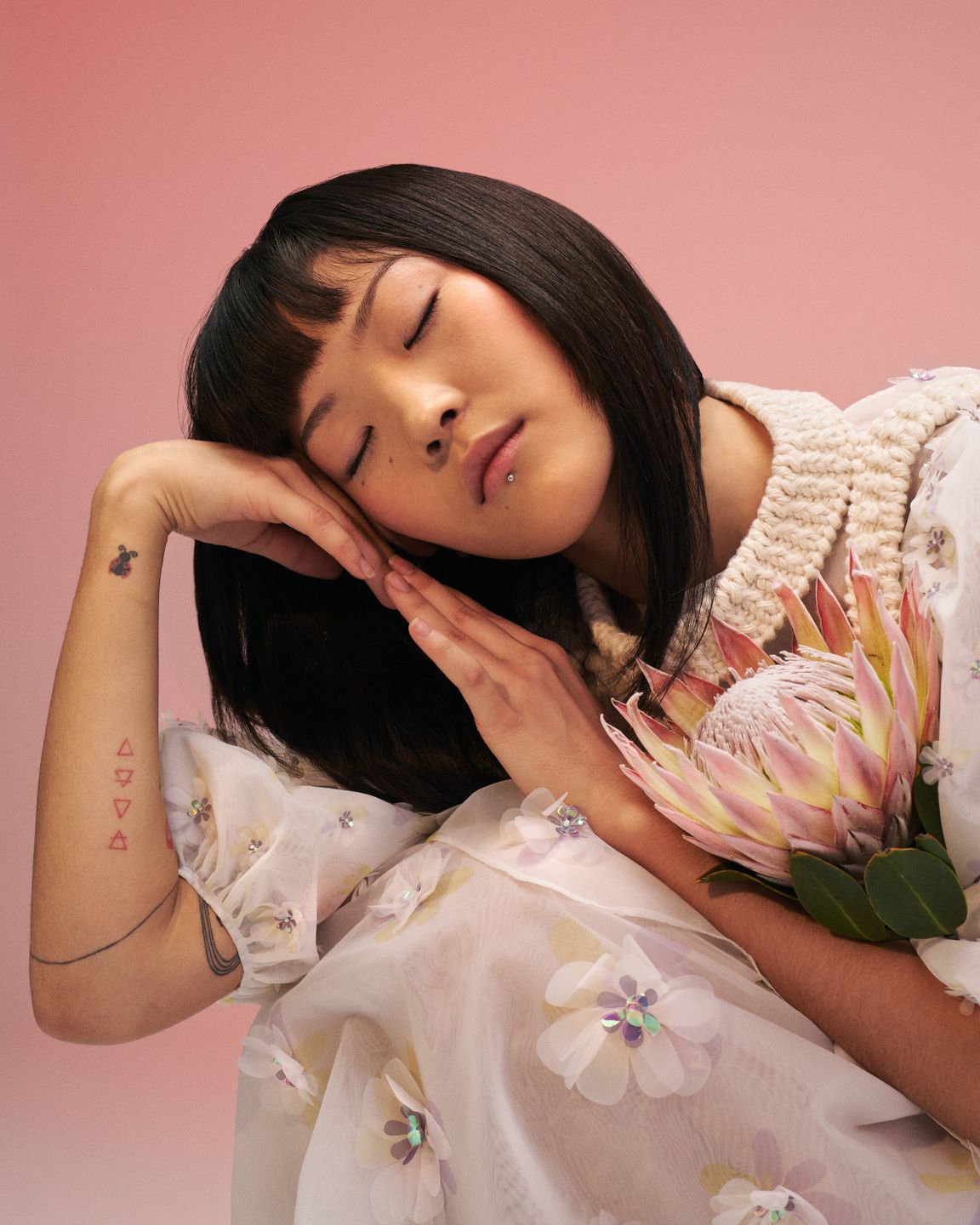 Model Anujin wears a Selina Rotach white dress, holding a Protea flower. SHe is resting her head on her hands and has her eyes closed. Hair and makeup by Denise Dedich.