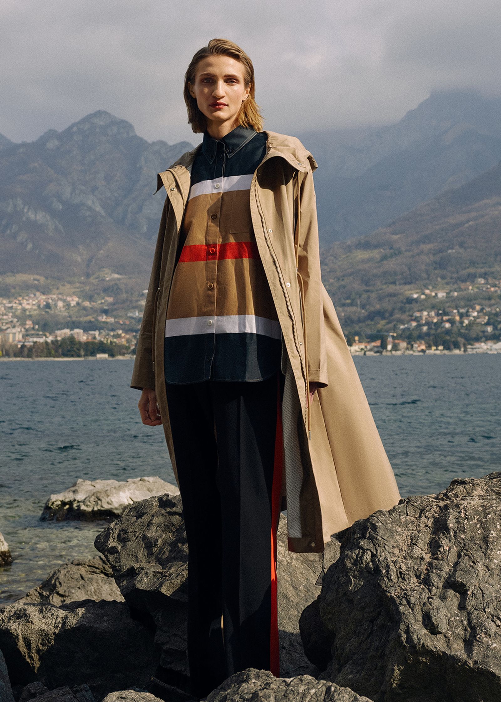 a woman in a trench coat standing on rocks next to a lake