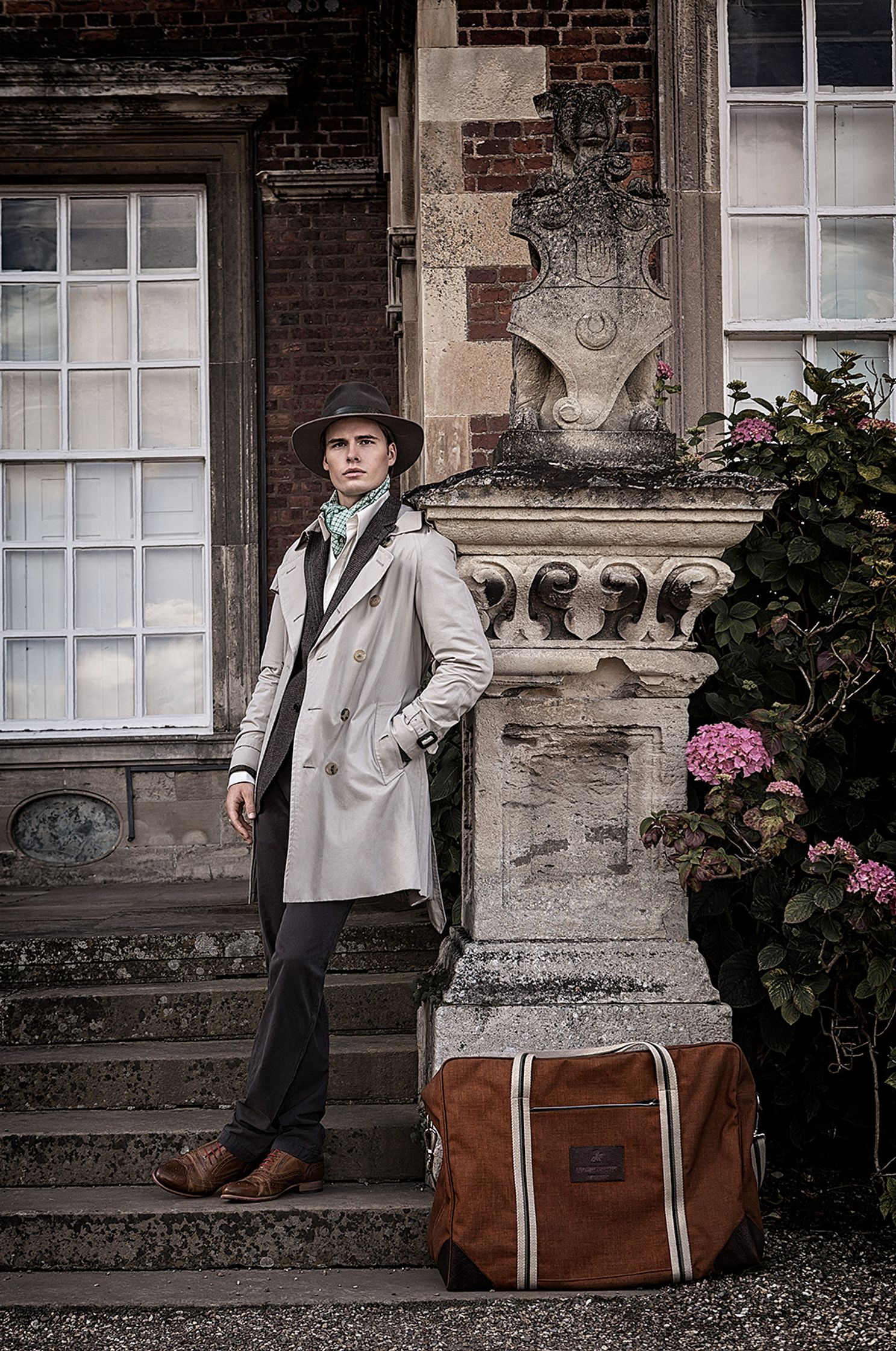 a man wearing a hat and coat standing next to a suitcase creative post production