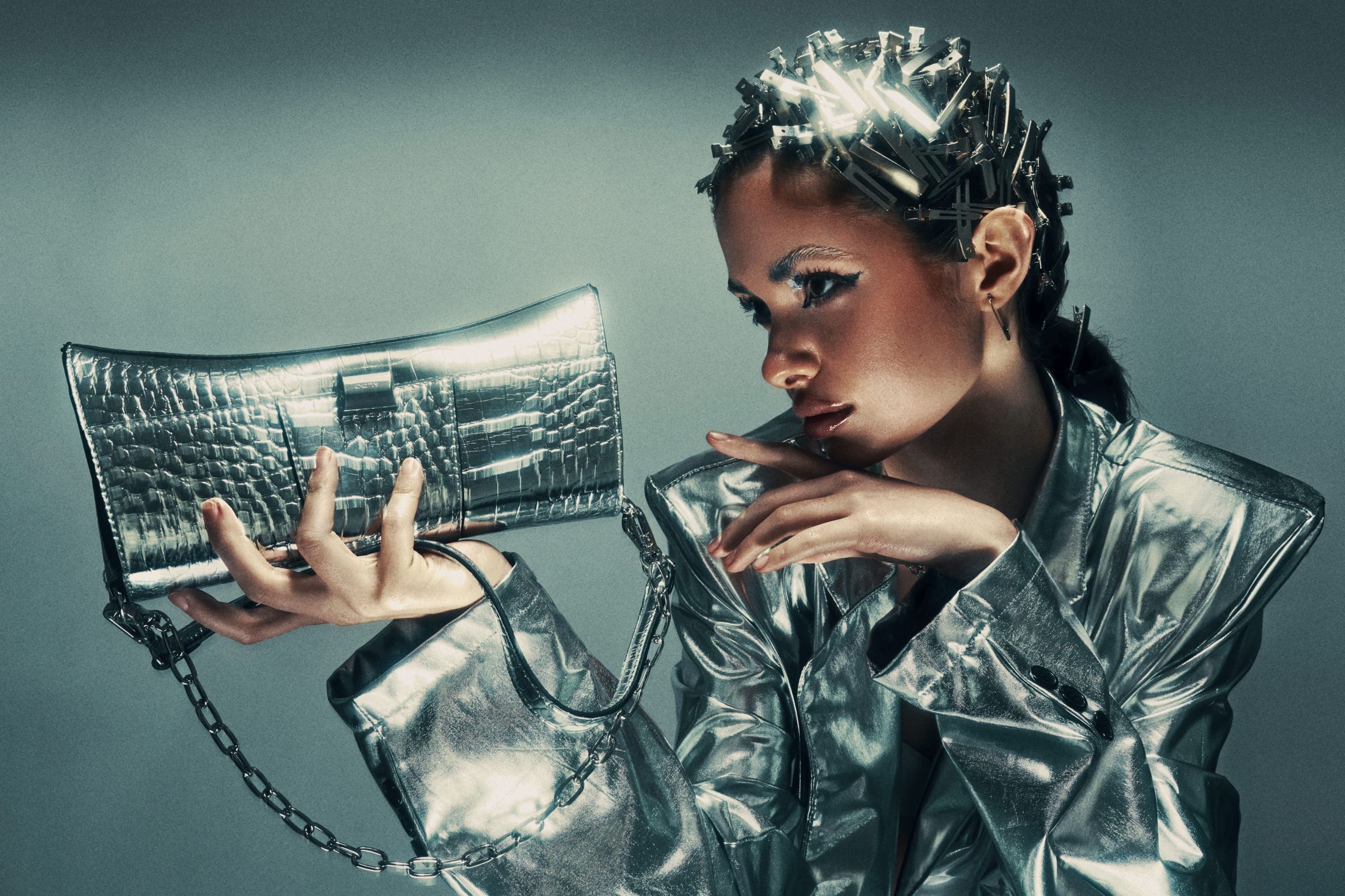 Dramatic close-up of Monique Bourscheid with bold makeup and silver hair accessories, resting her chin on her hand while holding a metallic handbag 