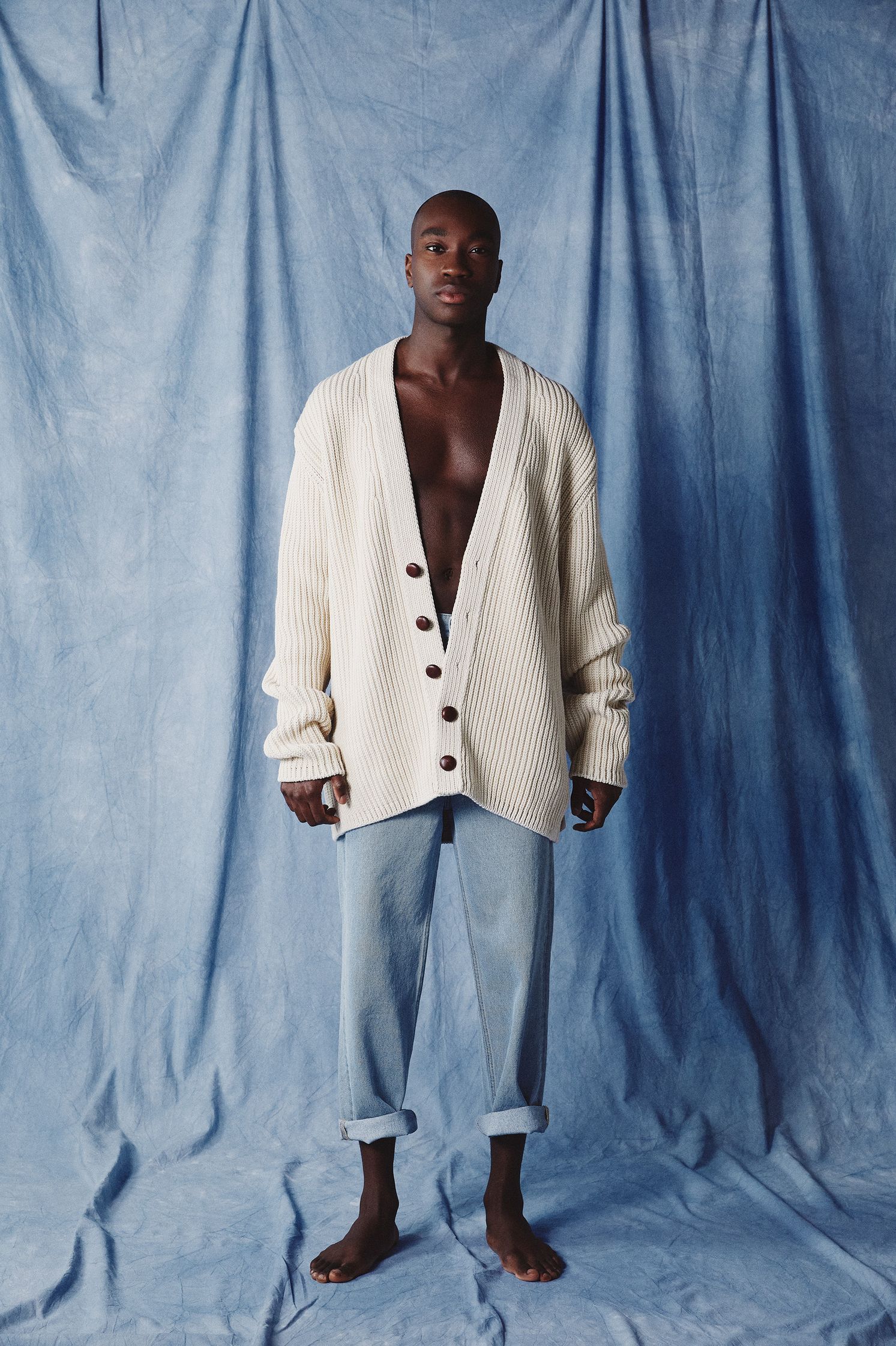 a man in a white cardigan and jeans standing in front of a blue backdrop