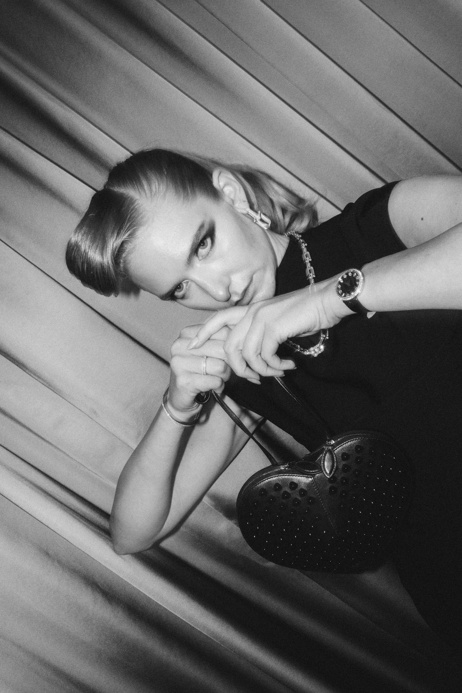 A black and white photo of model Jasmin Brunner, posing in a black Maison Alaia dress. She wears Tiffany earrings, necklace and watch. She looks straight into the camera. Her foundation is dewy, her eye makeup is smokey and glam. Makeup by Denise Dedich.