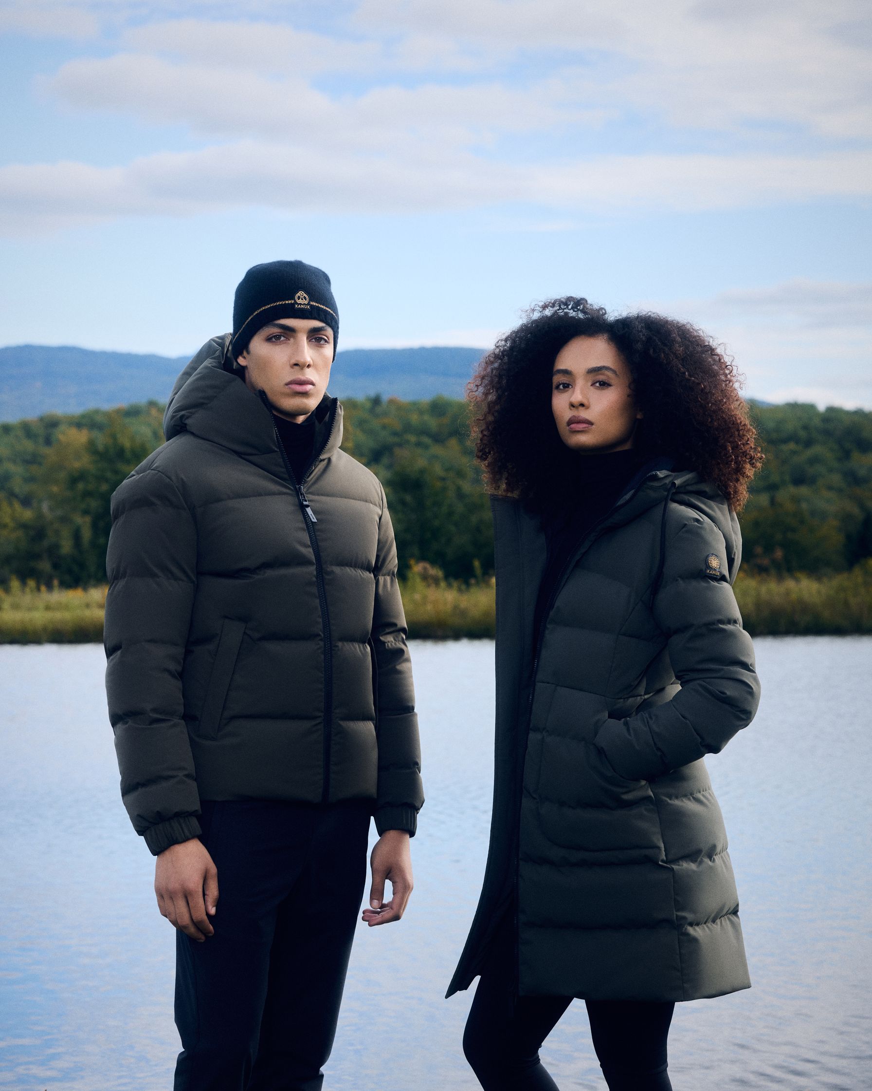 a man and woman standing next to a lake wearing down jackets