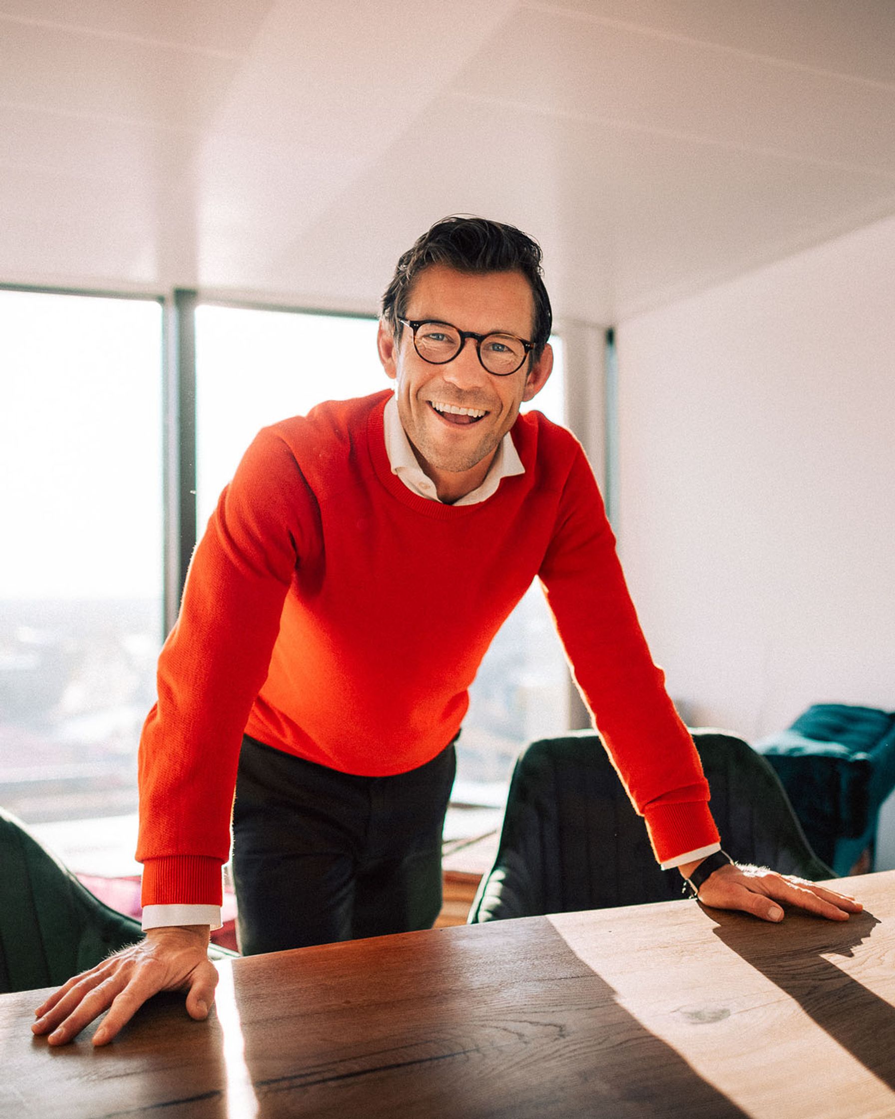 a man wearing glasses and a red sweater is leaning against a desk