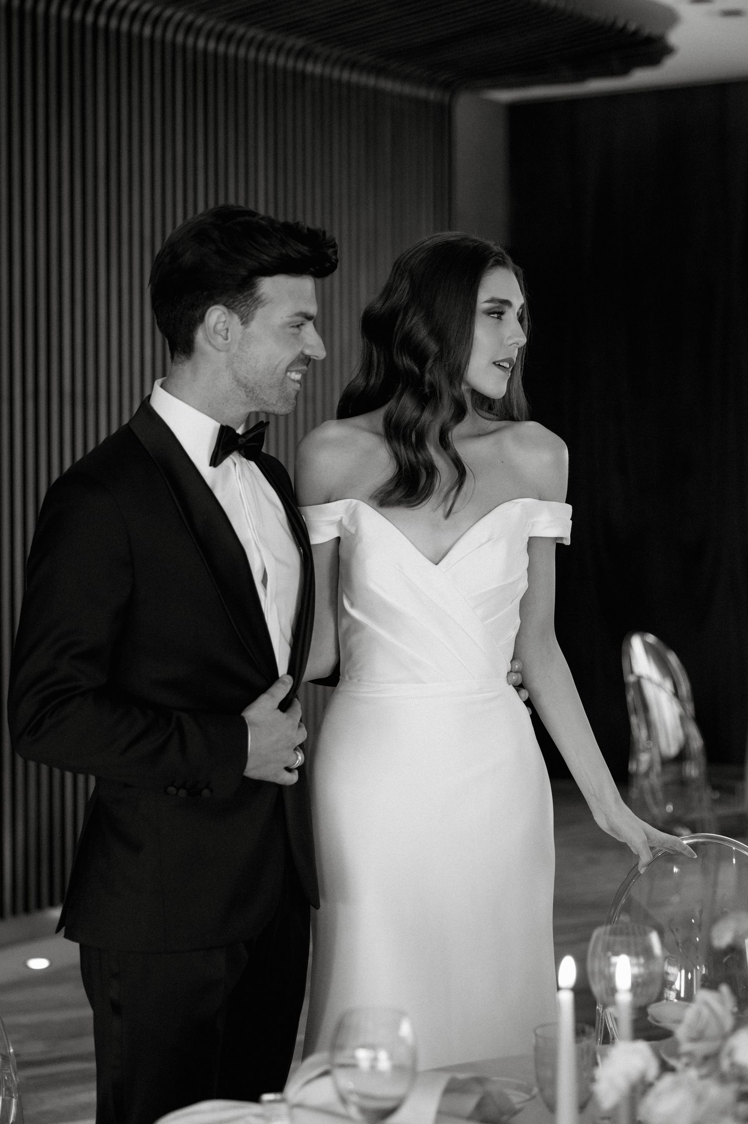 Vivienne Oesch and Vasilis Zorba posing as bride and groom at a wedding table. She wears a a white, off-the-shoulders dress, he wears a tuxedo. They look away from the camera. Bridal makeup in Zürich. Hair and makeup by Denise Dedich.