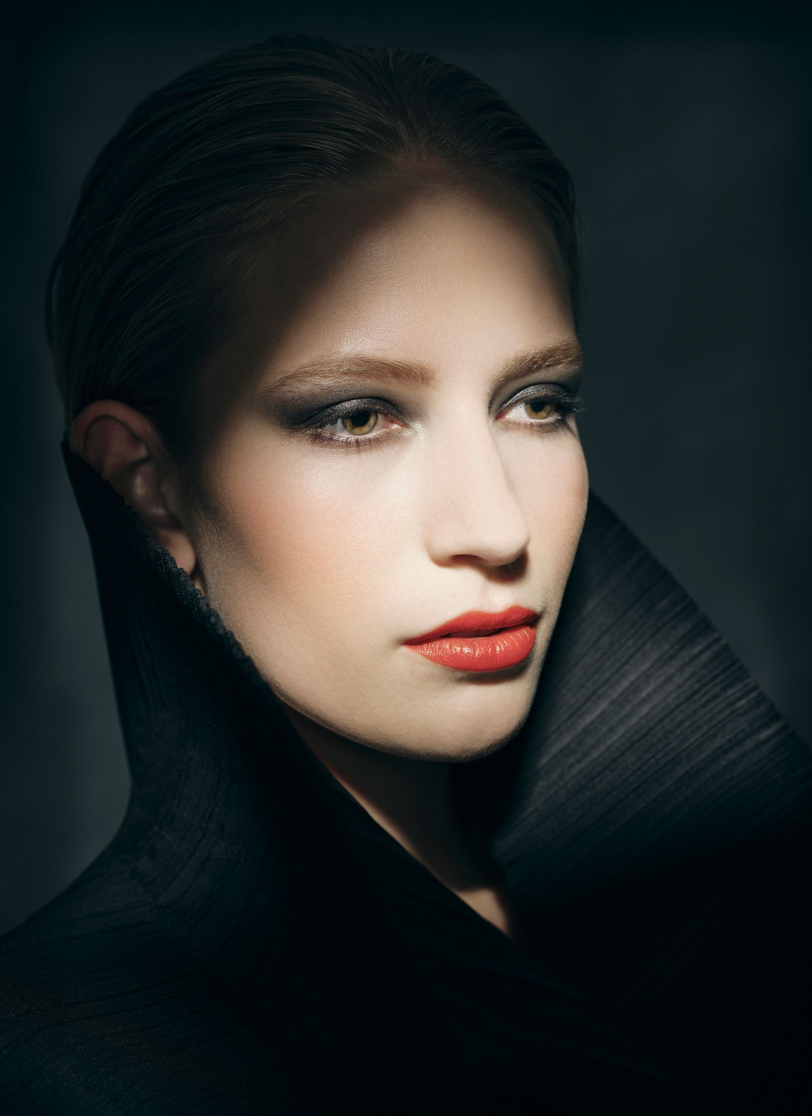 Close-up of model Joy Emmenegger in a black coat posing in front of a dark background. Red lipstick, glam makeup. Hair and makeup by Denise Dedich.