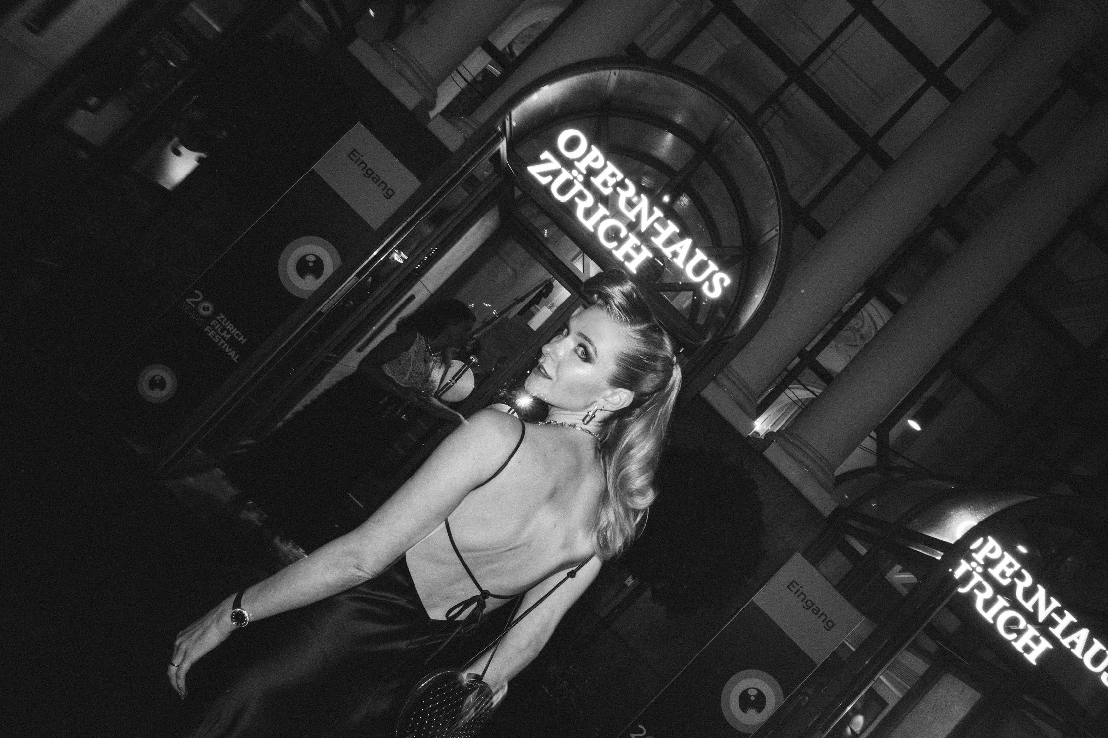 A black and white photo of model Jasmin Brunner, posing in a black dress in front of the Zurich Opernhaus during the Zurich Film Festival. She wears Tiffany earrings, necklace and watch. Her foundation is dewy, her eye makeup is smokey and glam. Makeup by Denise Dedich.