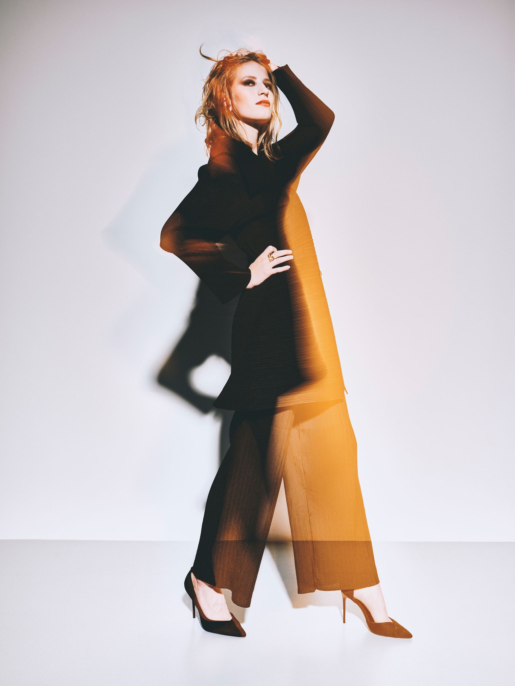 Model Joy Emmenegger  in a black outfit posing in front of a white wall, the image is a bit blurry. Red lipstick, glam makeup. Hair and makeup by Denise Dedich.