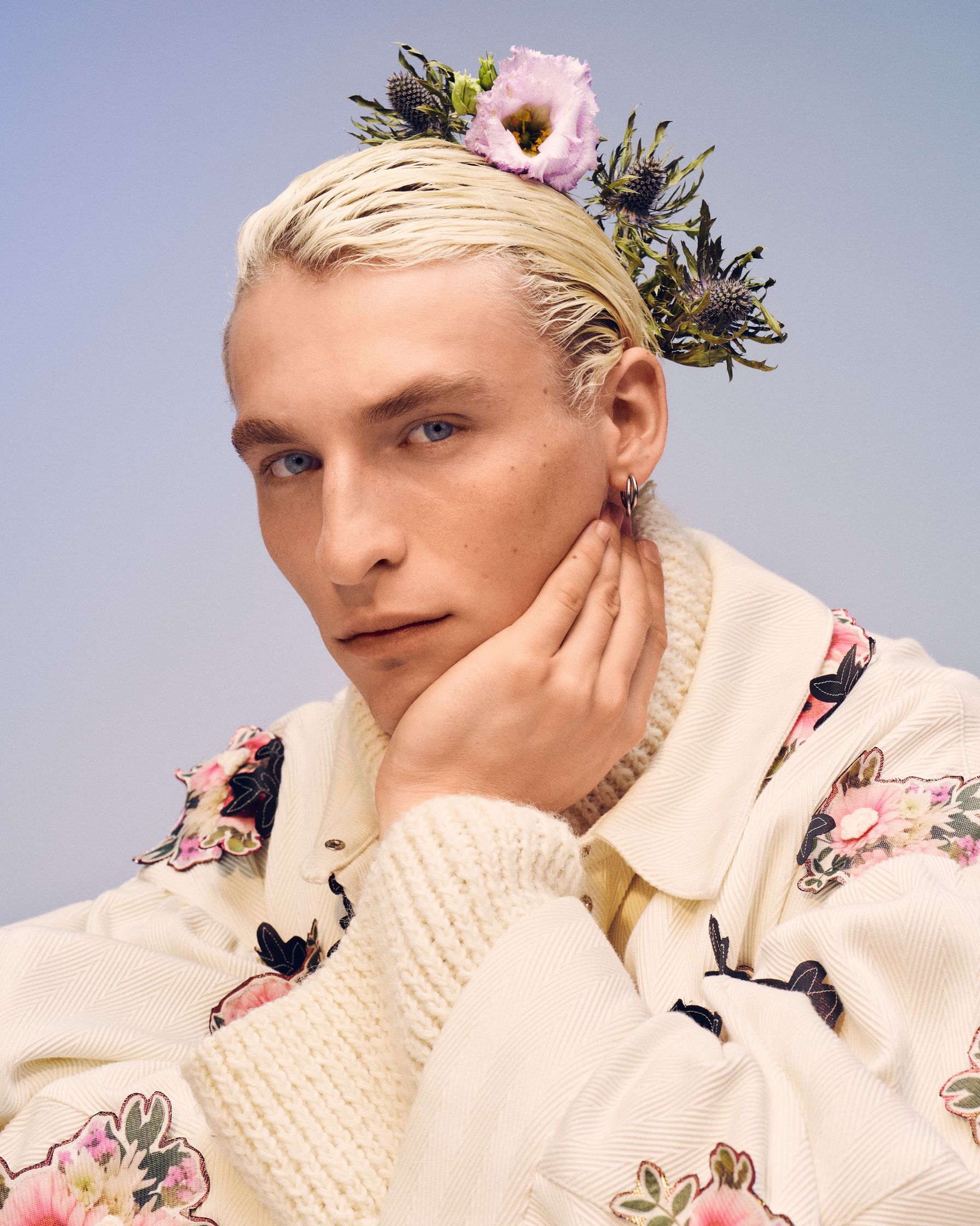 Model Livio Michel is wearing a Selina Rotach white jacket and has flowers on his head. His hair is sleeked back. Grooming by Denise Dedich.