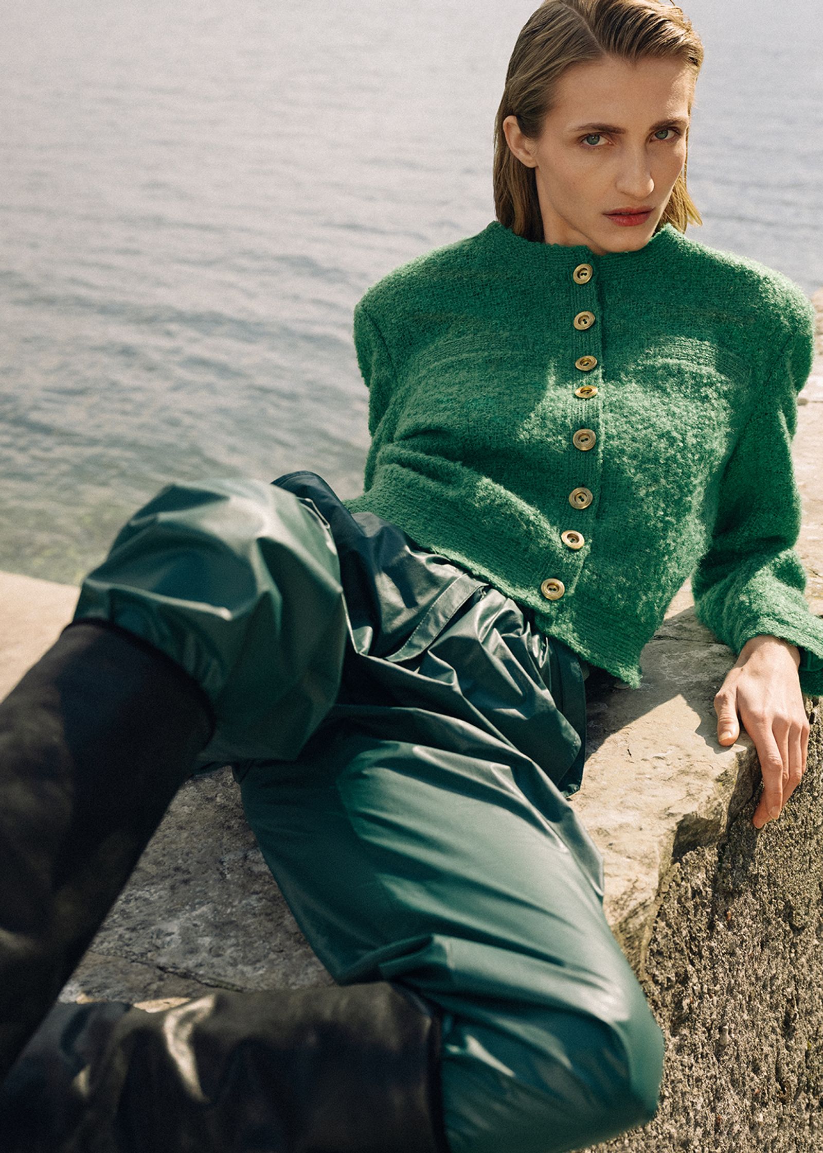 a woman in a green sweater and leather pants leaning against a wall